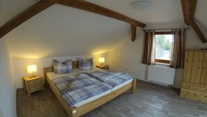 Ein Schlafzimmer mit schrägen Wänden, Holzbalken, einem Bett mit kariertem Bezug, Nachttischlampen und einem Fenster mit Vorhängen.
