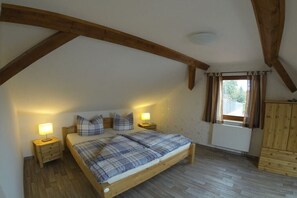 Ett sovrum med ett sluttande tak, synliga träbjälkar, en säng med ett rutigt överkast, sänglampor och ett fönster med gardiner.