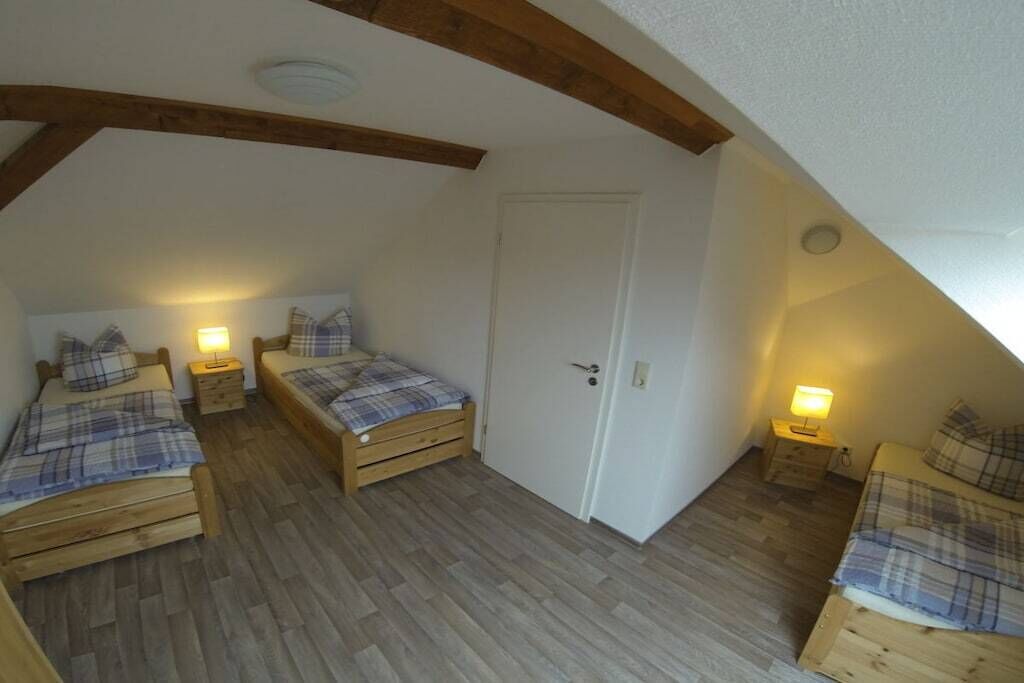 A small attic bedroom with three wooden beds, each with a matching headboard and footboard. There are bedside tables with lamps, and the room has a sloped ceiling with exposed wooden beams.