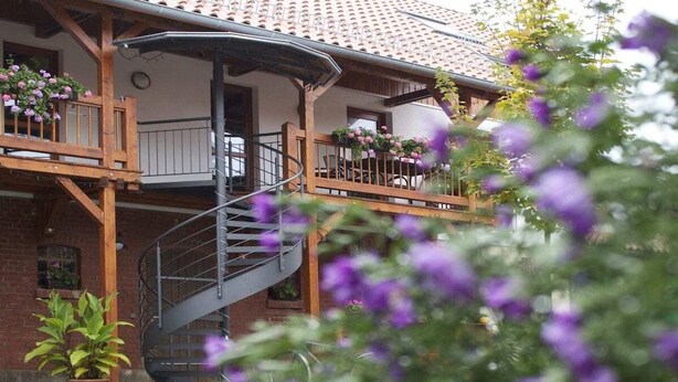 Ein zweigeschossiges Gebäude mit einer Wendeltreppe, die zu einem Balkon führt, umgeben von Blumentöpfen und Blumen.