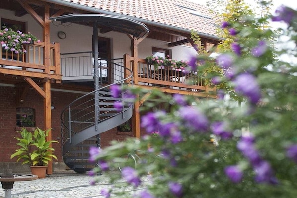 En tvåvåningsbyggnad med en spiraltrappa som leder upp till en balkong, omgiven av planterade växter och blommor.