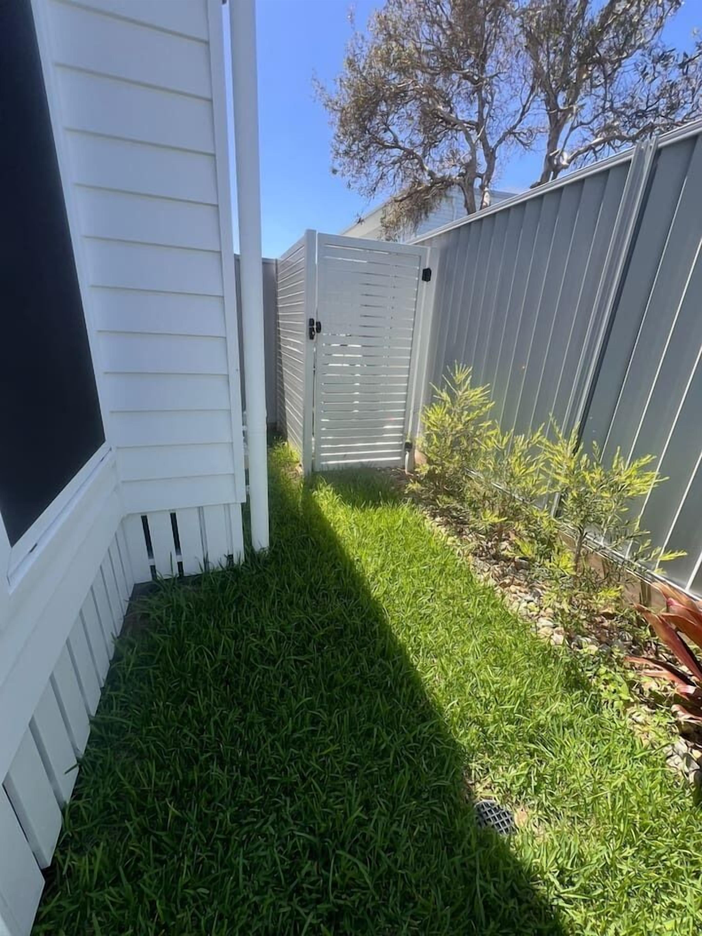 A narrow grassy pathway flanked by white and gray fences, leading to a building with a dark window.