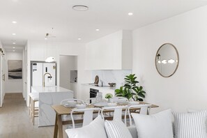 A modern kitchen with white cabinetry, a central island, and a dining area with a wooden table and white chairs. - Your Buddina Escape:Swim, Sip & Unwind by the Pool (Buddina)