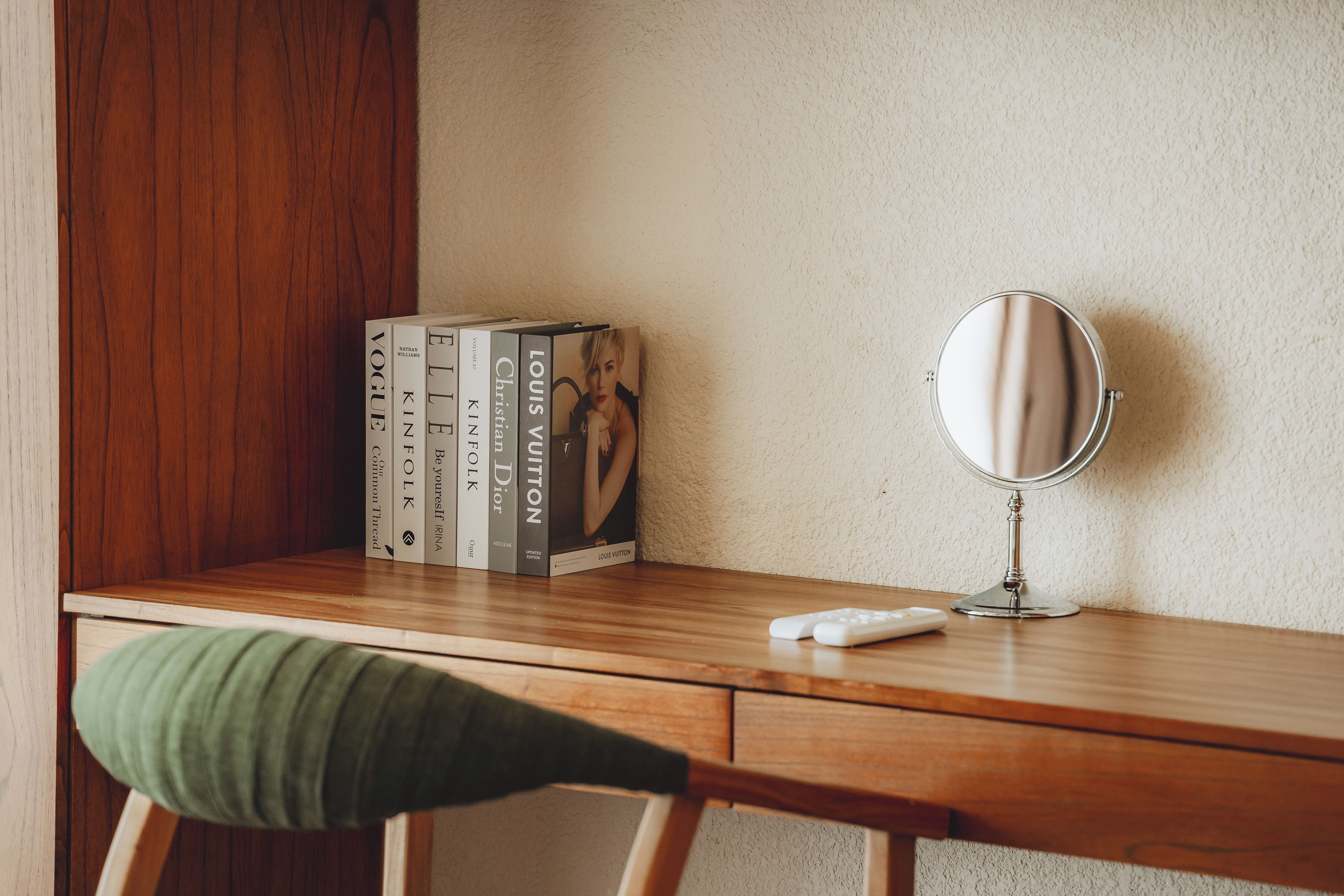 Uma mesa de madeira com um espelho, uma cadeira verde e uma pilha de livros.