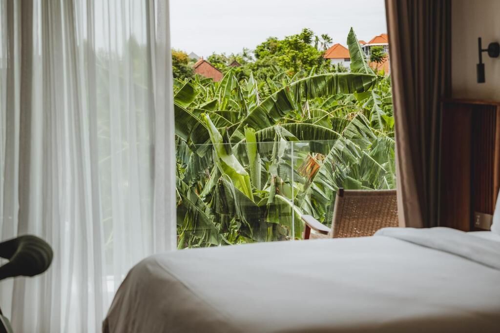 A hotel room with a bed, a chair, and a view of lush greenery and buildings outside.