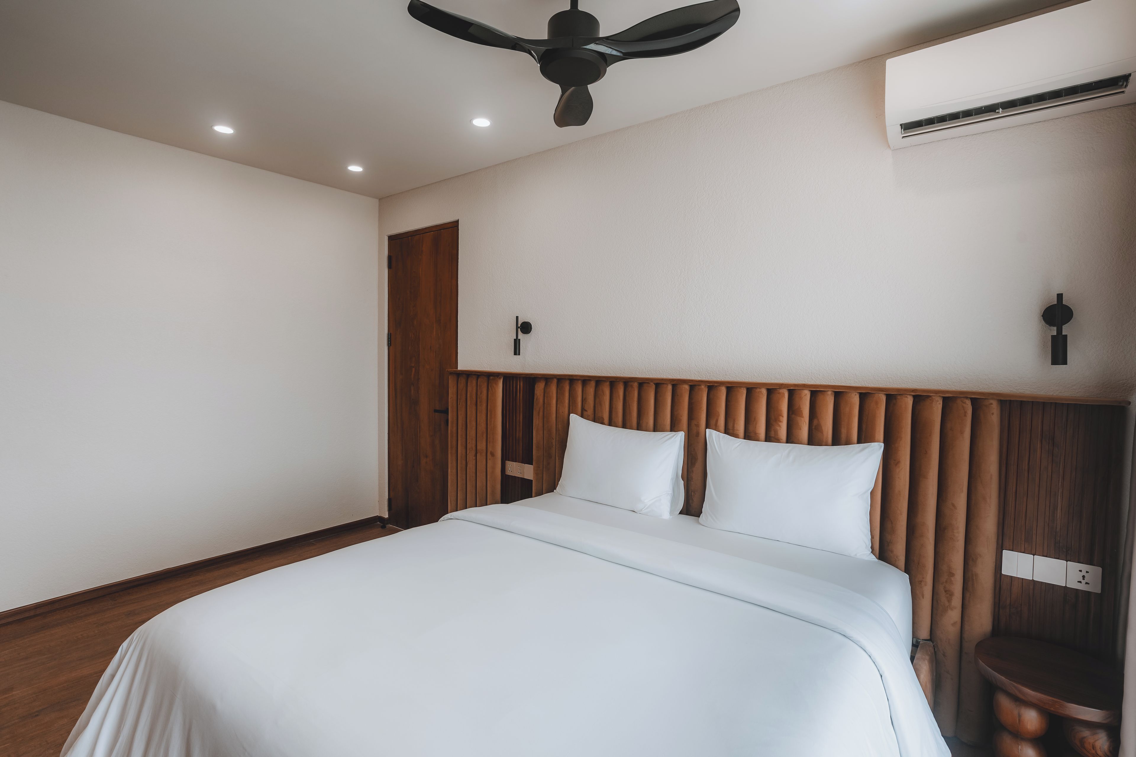 A hotel room with a wooden headboard, a bed with white bedding, a ceiling fan, recessed lighting, and an air conditioning unit.
