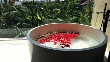 Una vasca da bagno ampia e rotonda piena d'acqua e petali di fiori rossi, posizionata su un balcone con vista su una zona residenziale e del verde.