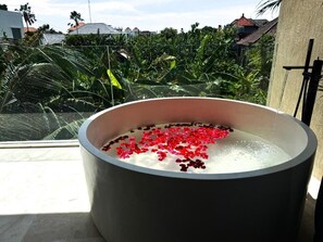 A large, round bathtub filled with water and red flower petals, placed on a balcony with a view of a residential area and greenery.