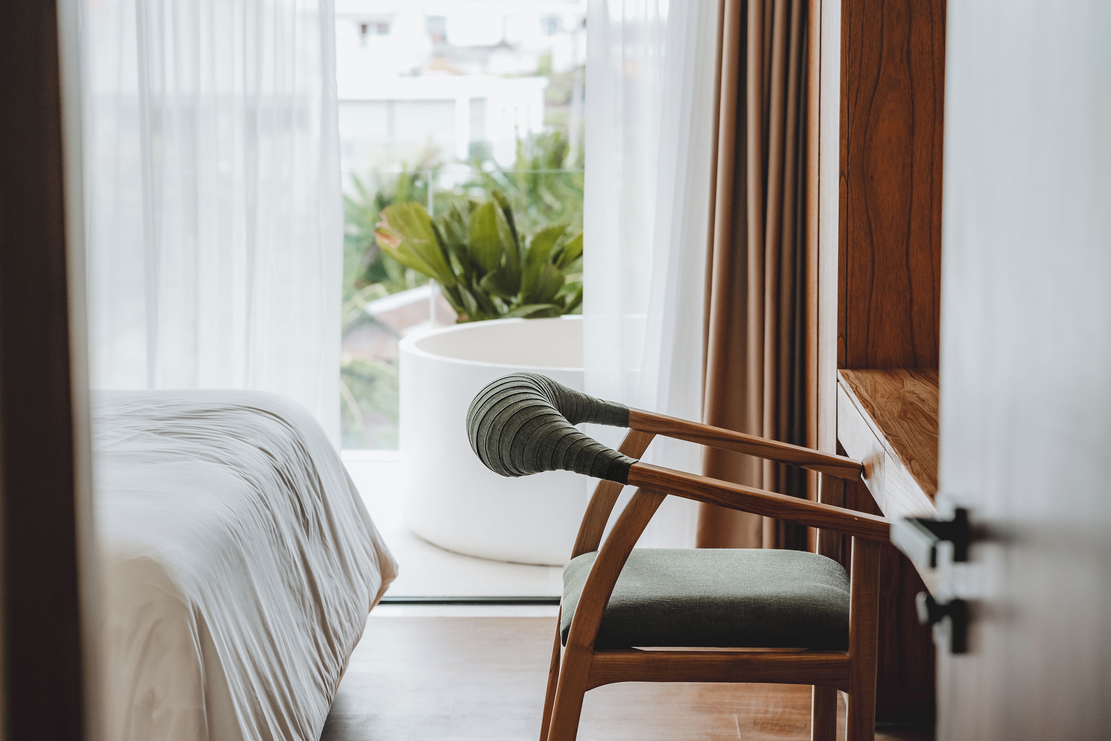 A room with a bed, a wooden chair with a cushion, a white round table, and a view of greenery outside.