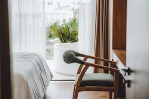 Una habitación con una cama, una silla de madera con cojín, una mesa redonda blanca y vistas a la vegetación exterior.