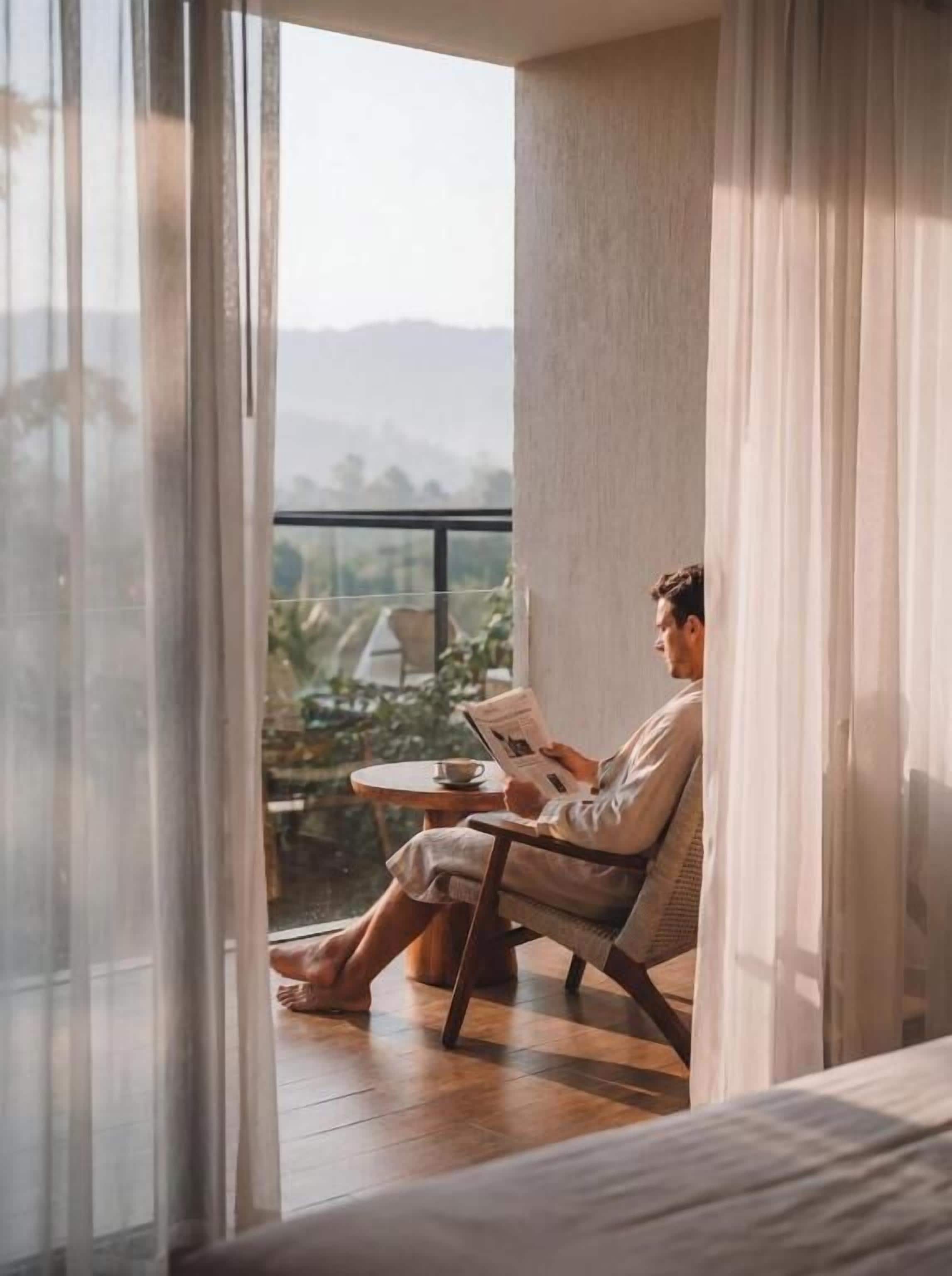 Una persona sentada en una silla junto a una ventana con vistas a zonas verdes y colinas.
