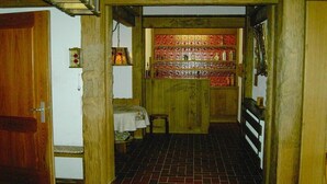 Een smalle gang met een houten deur aan de linkerkant, een klein tafeltje met een stoel, en een plank met flessen.