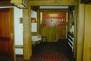 A narrow hallway with a wooden door on the left, a small table with a chair, and a shelf with bottles.