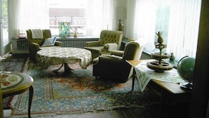Een kamer met een groot raam, een ronde tafel, twee fauteuils, een klein tafeltje met een wereldbol, en een kamerbreed tapijt met een patroon.