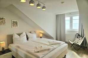 A bedroom with a bed, bedside lamps, a chair, and a window with curtains. - Beach oasis, sea breeze (Sierksdorf)