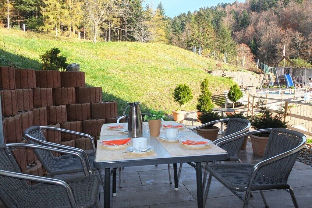 Une terrasse extérieure avec une table dressée pour deux personnes, entourée de chaises en osier, offrant une vue sur une aire de jeux et une colline verdoyante en arrière-plan.