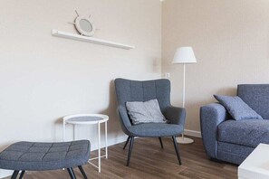 A modern living room with a grey armchair, a matching ottoman, and a blue sofa. A white floor lamp and a small round table with a white base are also visible. - House Bärbel Apartment No. 5 (Cuxhaven)