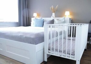 A white crib with a grey mattress, a white nightstand with a lamp, and a grey wall. - House Bärbel Apartment No. 6 (Cuxhaven)