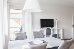 Un espace repas moderne comprenant une table, des chaises, un téléviseur posé sur un meuble blanc et une fenêtre avec des rideaux.