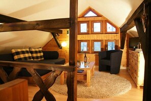 A cozy loft-style room with a sloped ceiling, wooden beams, a sofa with checkered cushions, a dark armchair, a wooden coffee table, and a television on a wooden dresser. - Forester's Lodge Apartment Tawny Owl (Burg)