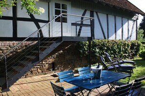 A traditional half-timbered house with a staircase leading to a balcony, outdoor seating with blue tables and chairs, and a stone wall foundation. - Holiday home with 3 bedrooms (Reichshof-Heischeid)