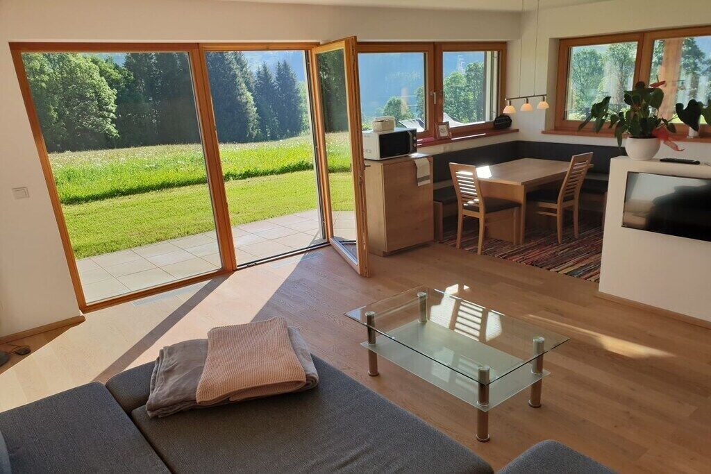 A modern living space with a glass dining table, a sofa, and a kitchen area with a view of a green landscape.