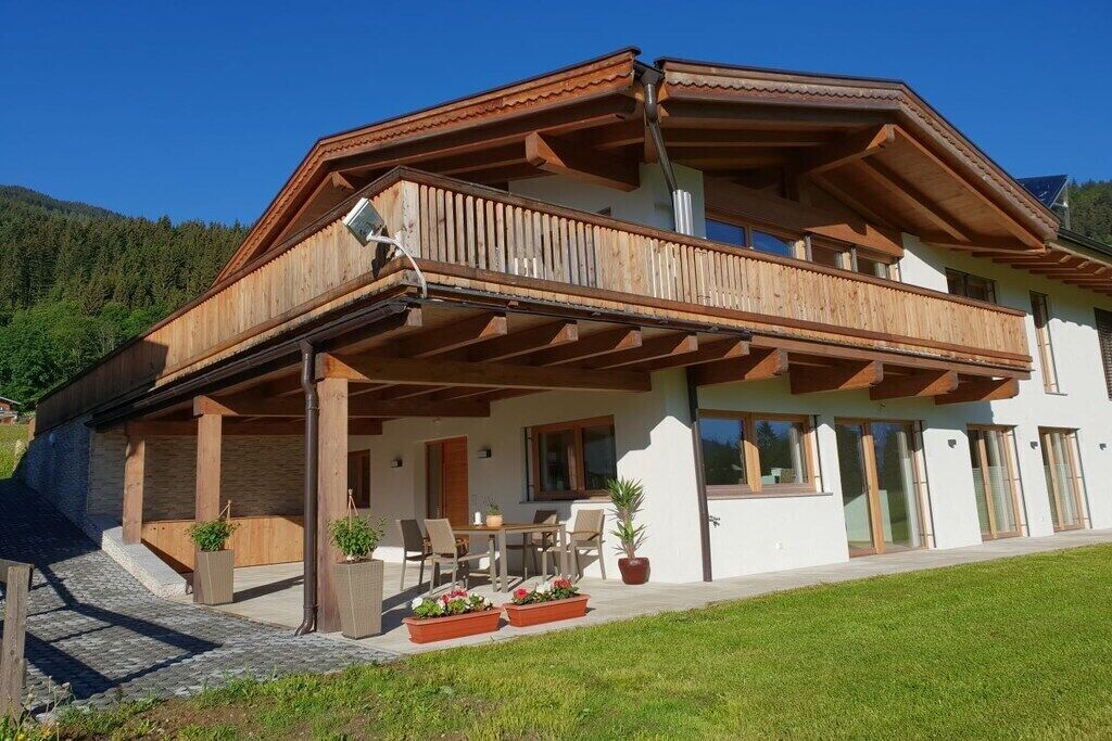 A two-story chalet with a wooden balcony, outdoor seating, and a mountain backdrop.