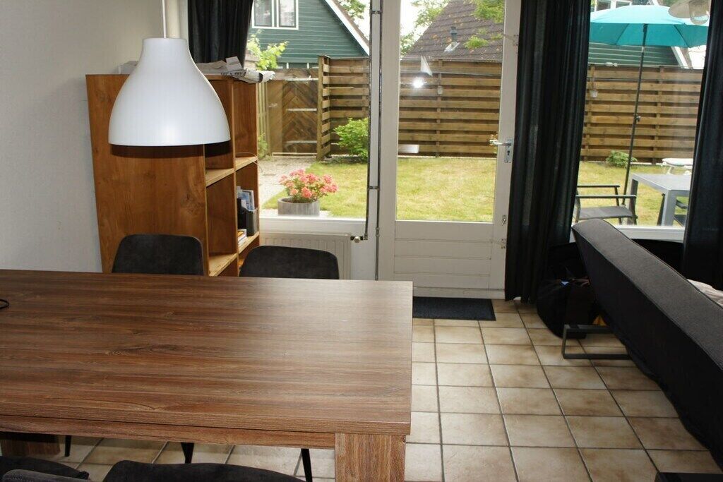 A room with a wooden table, chairs, a white lamp, a bookshelf, and a sliding glass door leading to a garden.