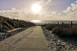 A paved pathway leading to a beach with a fence on one side and a grassy area on the other. - Holiday home with 3 bedrooms (Sint Maartensvlotbrug)