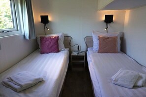 Ein Doppelzimmer mit zwei Einzelbetten, jeweils mit einem Kissen und einem gefalteten Handtuch. Es gibt Nachttische mit Lampen und eine kleine Ablagefläche.
