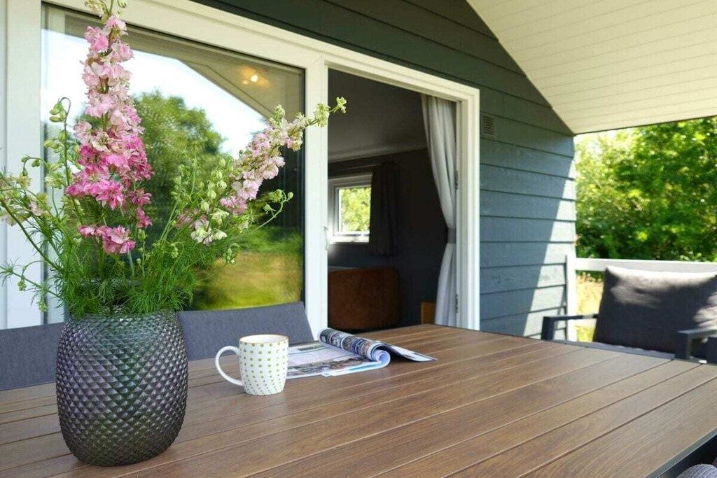 Une table en bois, sur laquelle sont posés un pot de fleurs, une tasse et un magazine, sur un balcon d’où l’on aperçoit une maison et de la verdure.