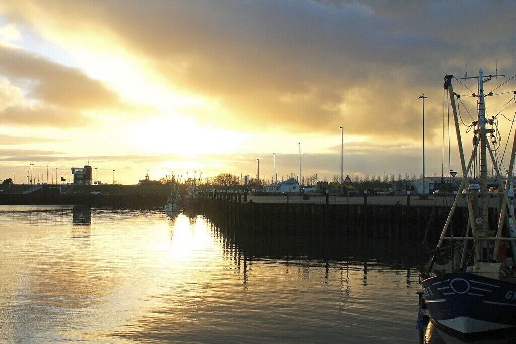Un port aux eaux calmes où le ciel se reflète, un quai avec un bateau, et un bâtiment situé au loin.
