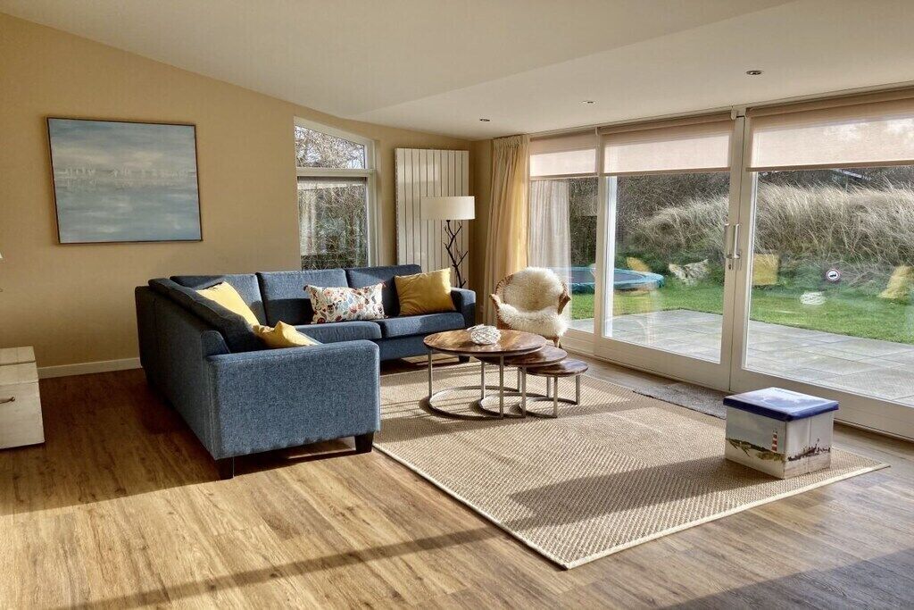 A living room with a sofa, coffee table, and a lamp. There is a window with curtains and a view of greenery outside.