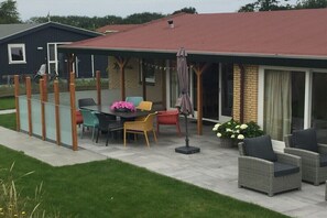 A covered patio area with a table and chairs, a potted plant, and a building with a red roof in the background. - Holiday apartment with 2 bedrooms (Ballum)