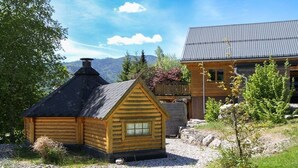 A wooden cabin with a sloped roof and a smaller adjacent structure, surrounded by a gravel area and greenery.