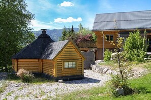 A wooden cabin with a sloped roof and a smaller adjacent structure, surrounded by a gravel area and greenery.