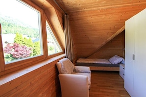 Habitació de fusta petita amb un llit individual, una cadira i una finestra gran que ofereix vistes a arbres i una casa.