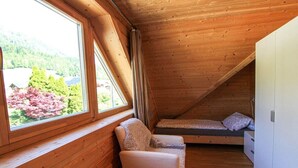 A small, wooden room with a single bed, a chair, and a large window offering a view of trees and a house.