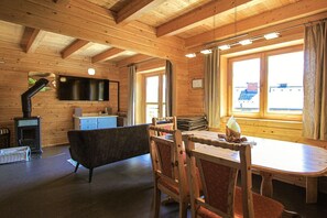 A wooden interior with a fireplace, a television, a dining table with chairs, and a view through large windows.