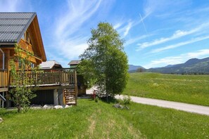 Une cabane en bois dotée d’une terrasse, entourée d’un champ verdoyant et avec des montagnes en arrière-plan.