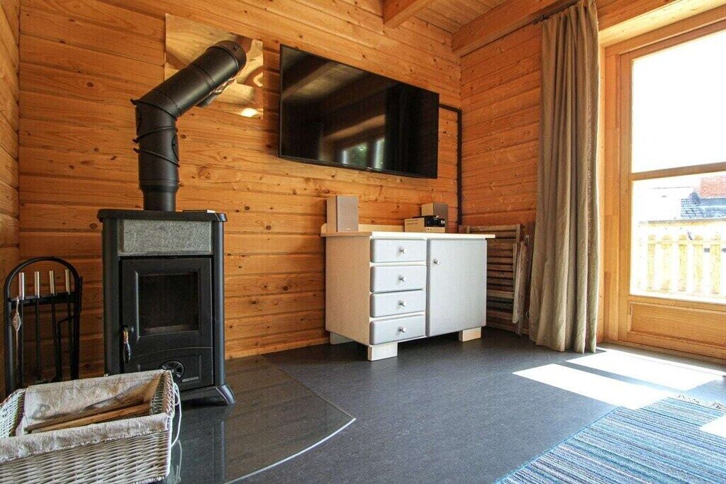 A room with a wood-burning stove, a television mounted on the wall, a white cabinet with drawers, and a window with curtains.