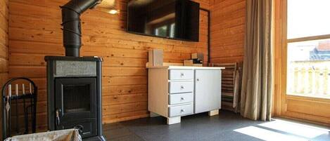 A room with a wood-burning stove, a television mounted on the wall, a white cabinet with drawers, and a window with curtains.
