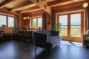 A wooden cabin interior with a dining area, a sofa, and large windows offering a view of the outdoors.