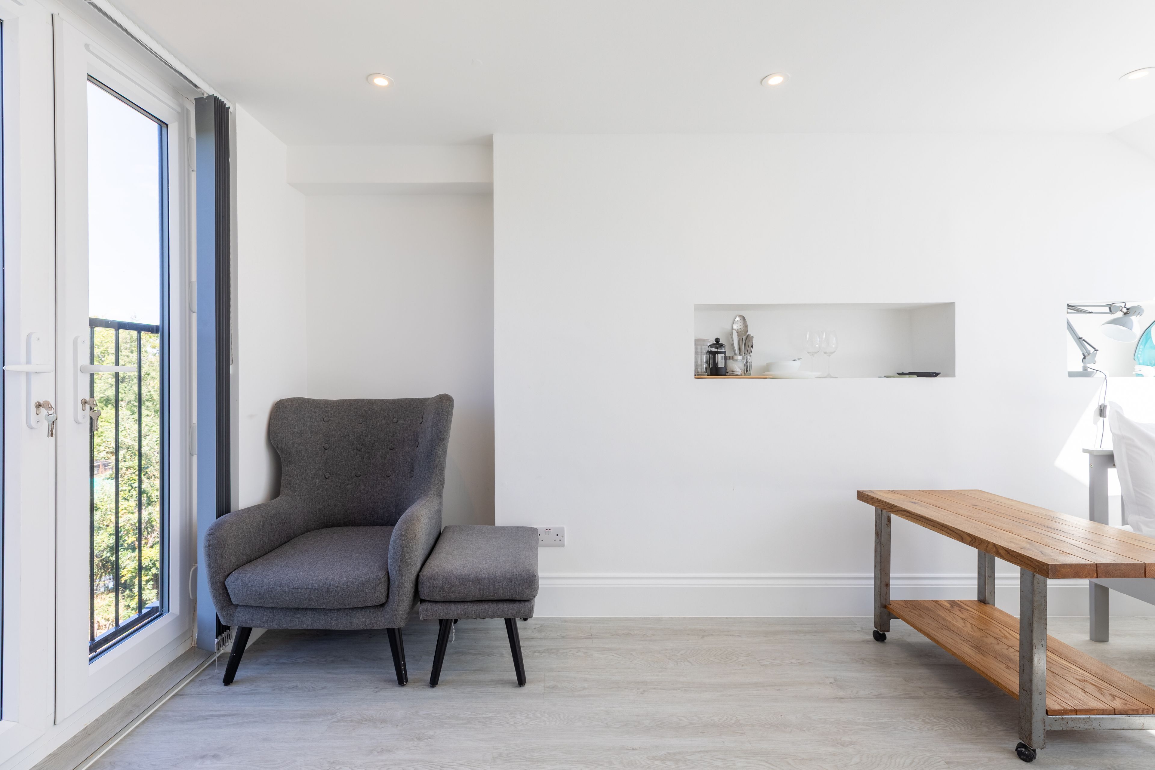 A modern room with a grey upholstered chair, a wooden table, and a white wall with a shelf.