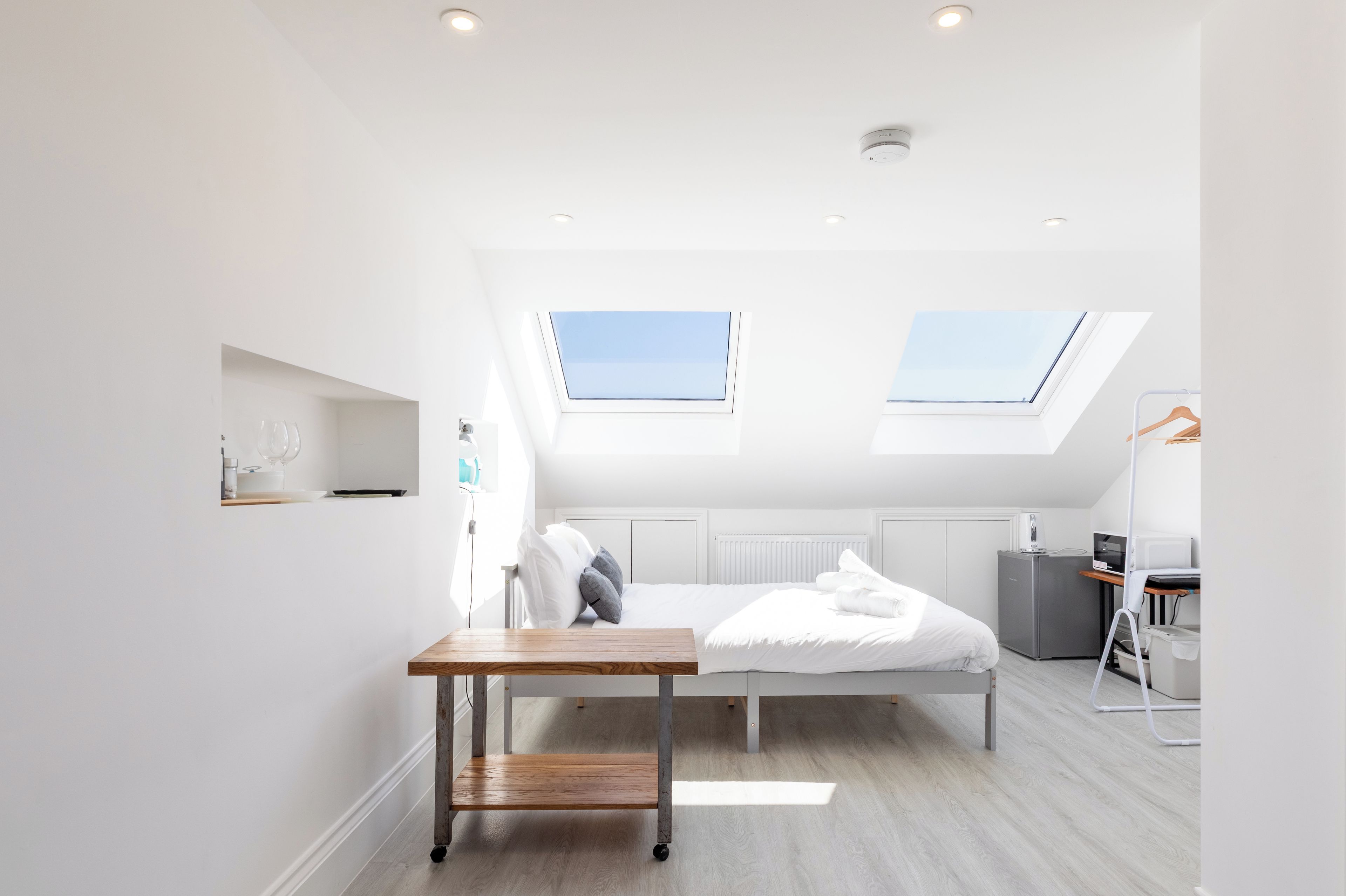A modern bedroom with a skylight, a bed with white bedding, a small table, and a compact kitchenette.