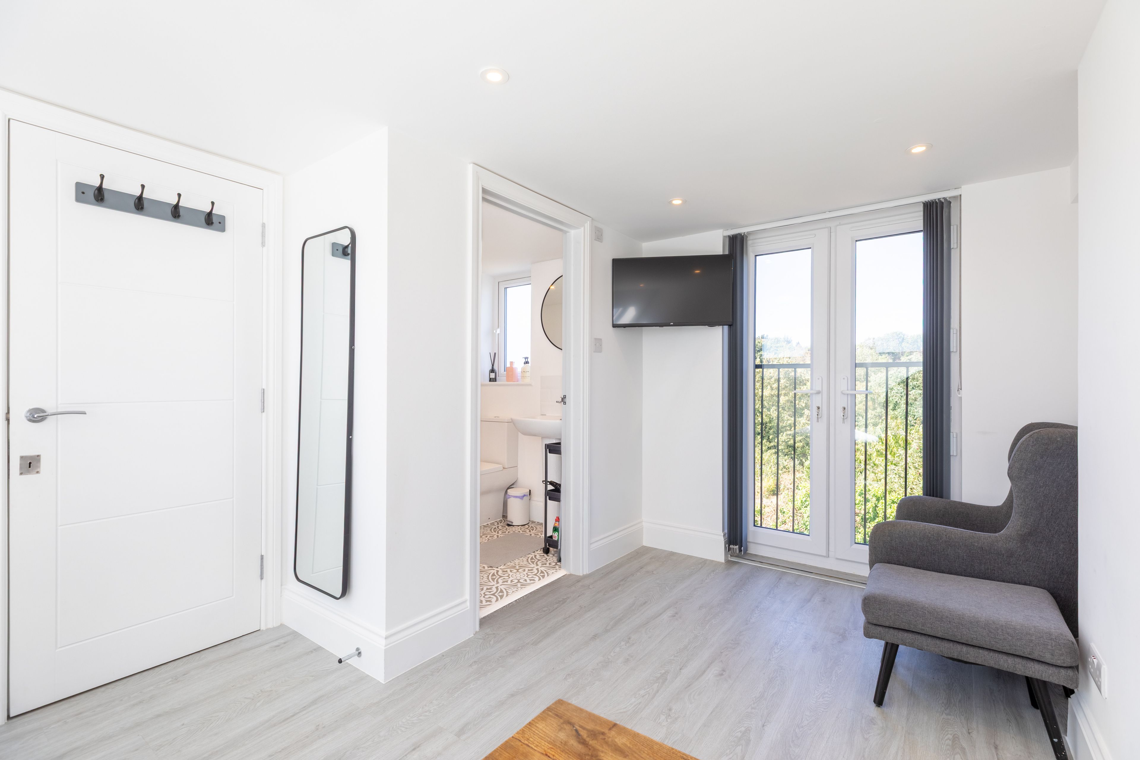 A modern living room with a grey sofa, a flat-screen TV mounted on the wall, and a balcony with a view.