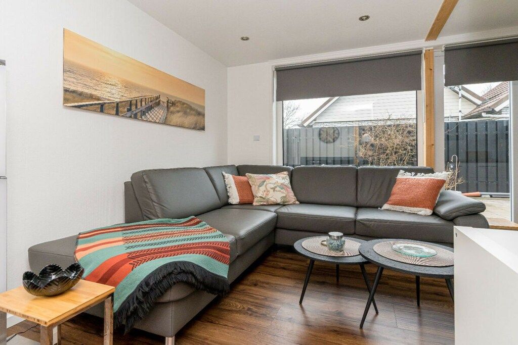Un salon moderne avec un canapé d’angle gris, une couverture tissée aux couleurs vives, une table d’appoint en bois et deux petites tables rondes.