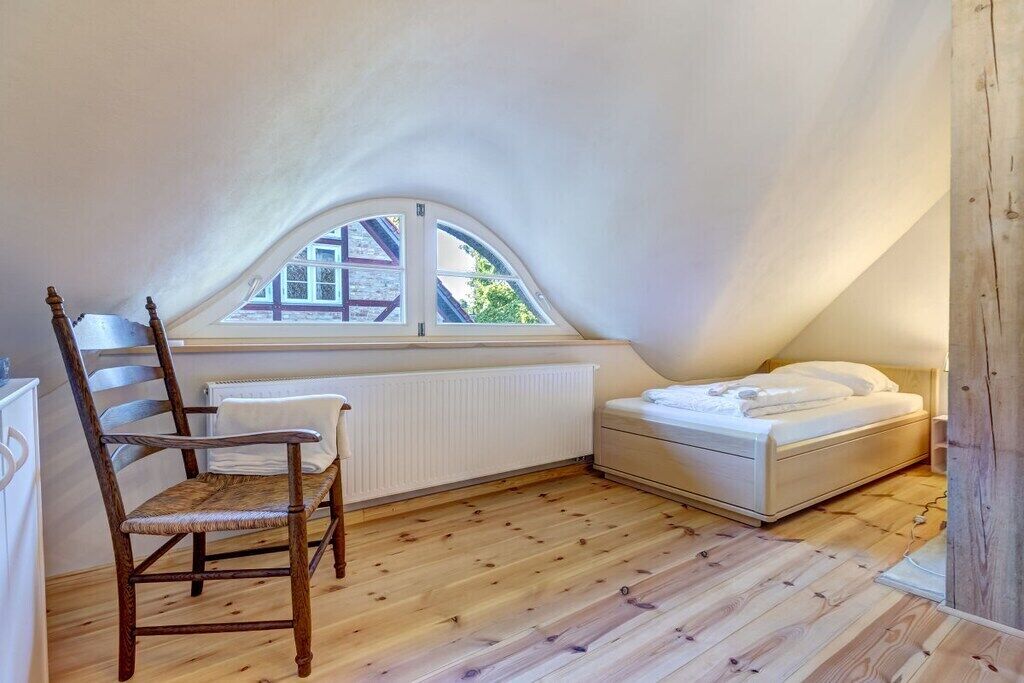 A small, cozy room with a sloped ceiling, a single bed, a wooden chair, and a window with a view of a building outside.