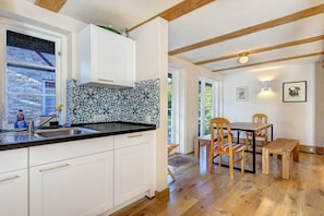 A kitchen with white cabinets, a black countertop, and a patterned backsplash. There is a dining area with wooden furniture and a window with a view of greenery. - Green Cottage by the Swan Pond (Lassan)