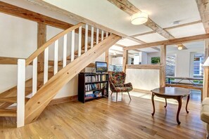 A wooden staircase with white balusters, a bookshelf, a floral armchair, and a round table in a room with wooden flooring and large windows. - Green Cottage by the Swan Pond (Lassan)
